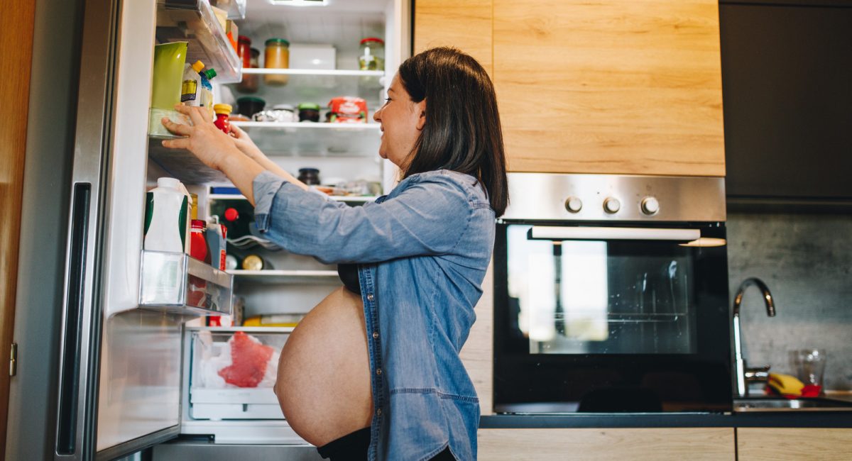 Lovely and beautiful pregnant woman, and a mother to be, standing in front of a open refrigerator, having pregnancy cravings.